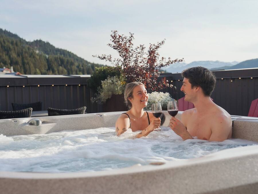 A hot tub with two people holding wine glasses, surrounded by wicker chairs and a wooden fence.