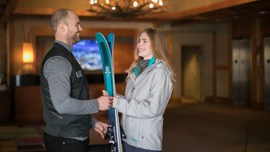 A man and a woman holding skis in a room with a chandelier and a television in the background. - Sundial Hotel (Whistler)