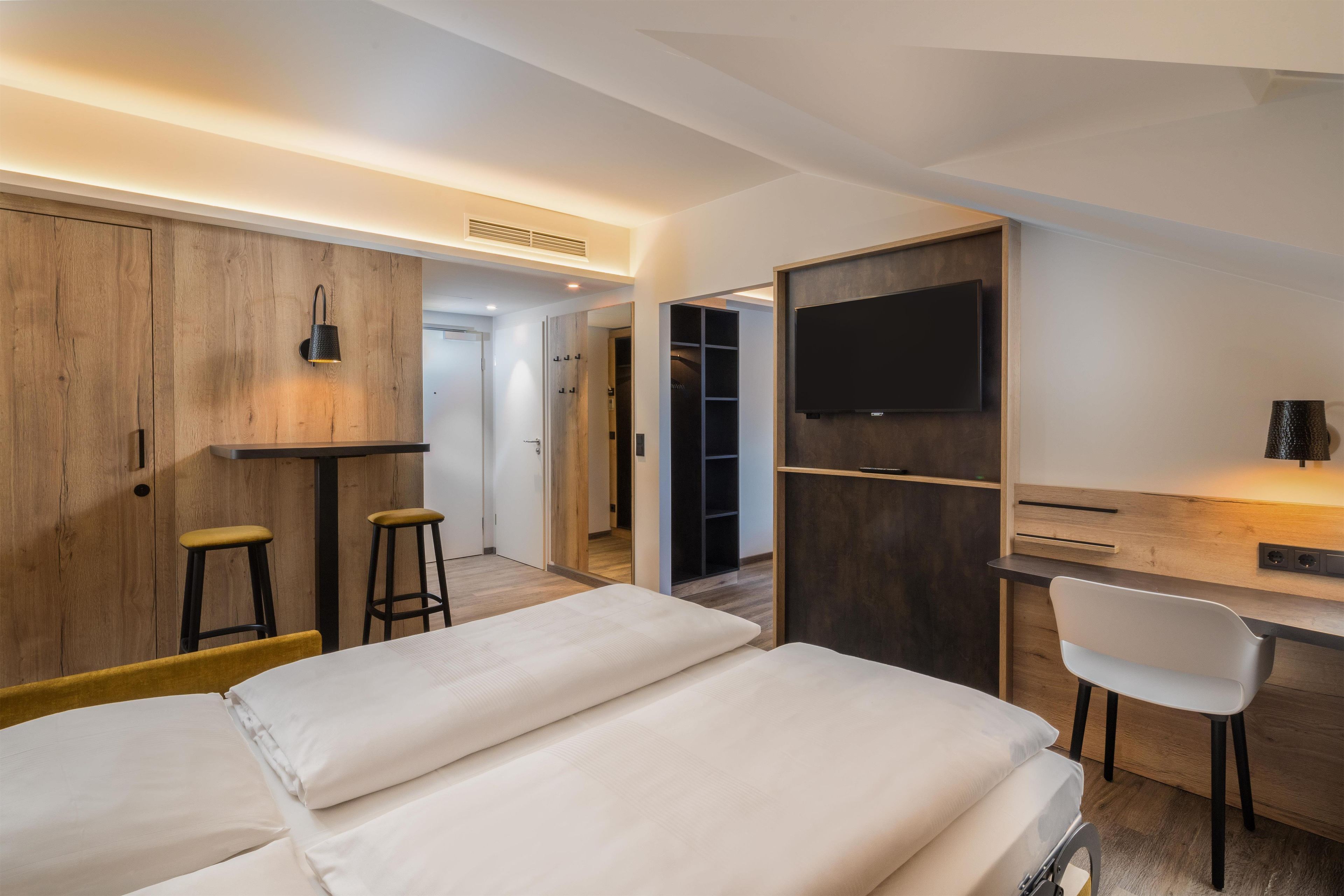 A modern hotel room with a bed, a desk with a chair, a TV, and a wardrobe.