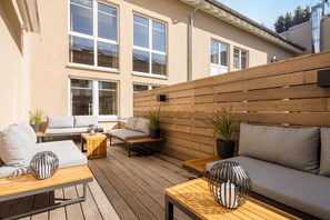 Een terras buiten met een houten dek, zithoek met banken en tafels, en een houten omheining.