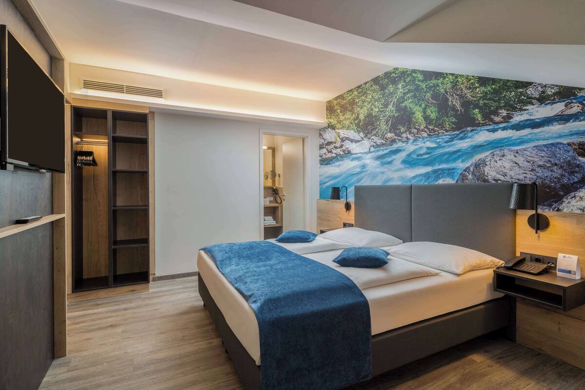 Een moderne hotelkamer met een groot bed, een kledingkast, een bureau en een schilderij van een rivier.