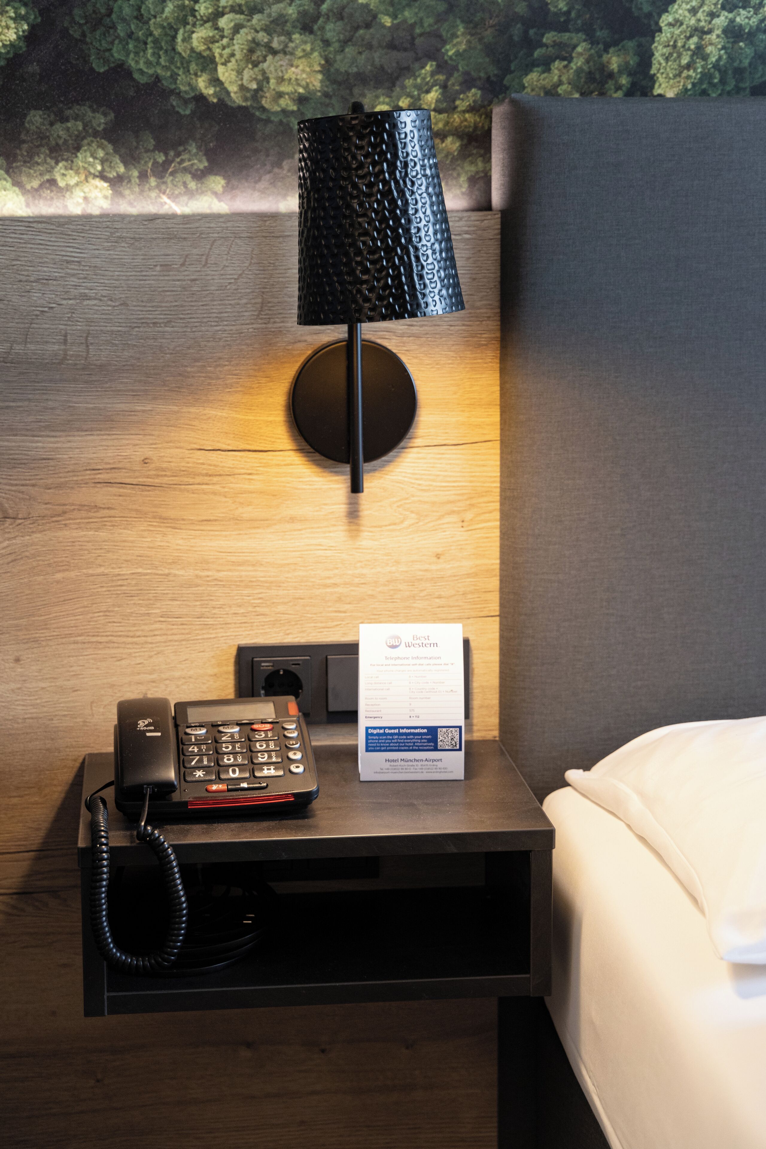Habitación de hotel con una mesita de noche de madera, un teléfono, una lámpara y un cartel con el nombre del hotel.