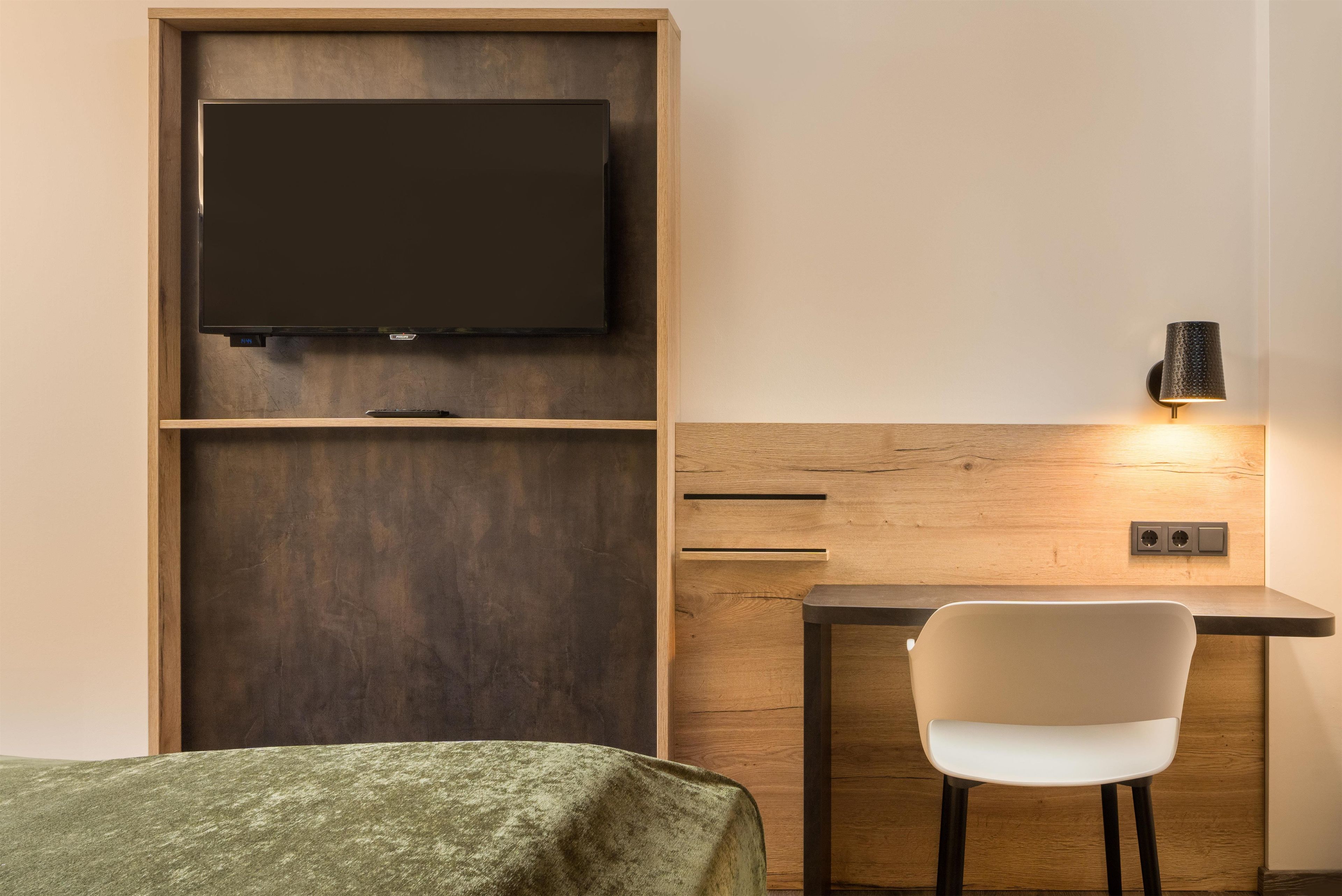 A hotel room with a bed, a desk with a chair, a television mounted on a wooden panel, and a wall-mounted lamp.
