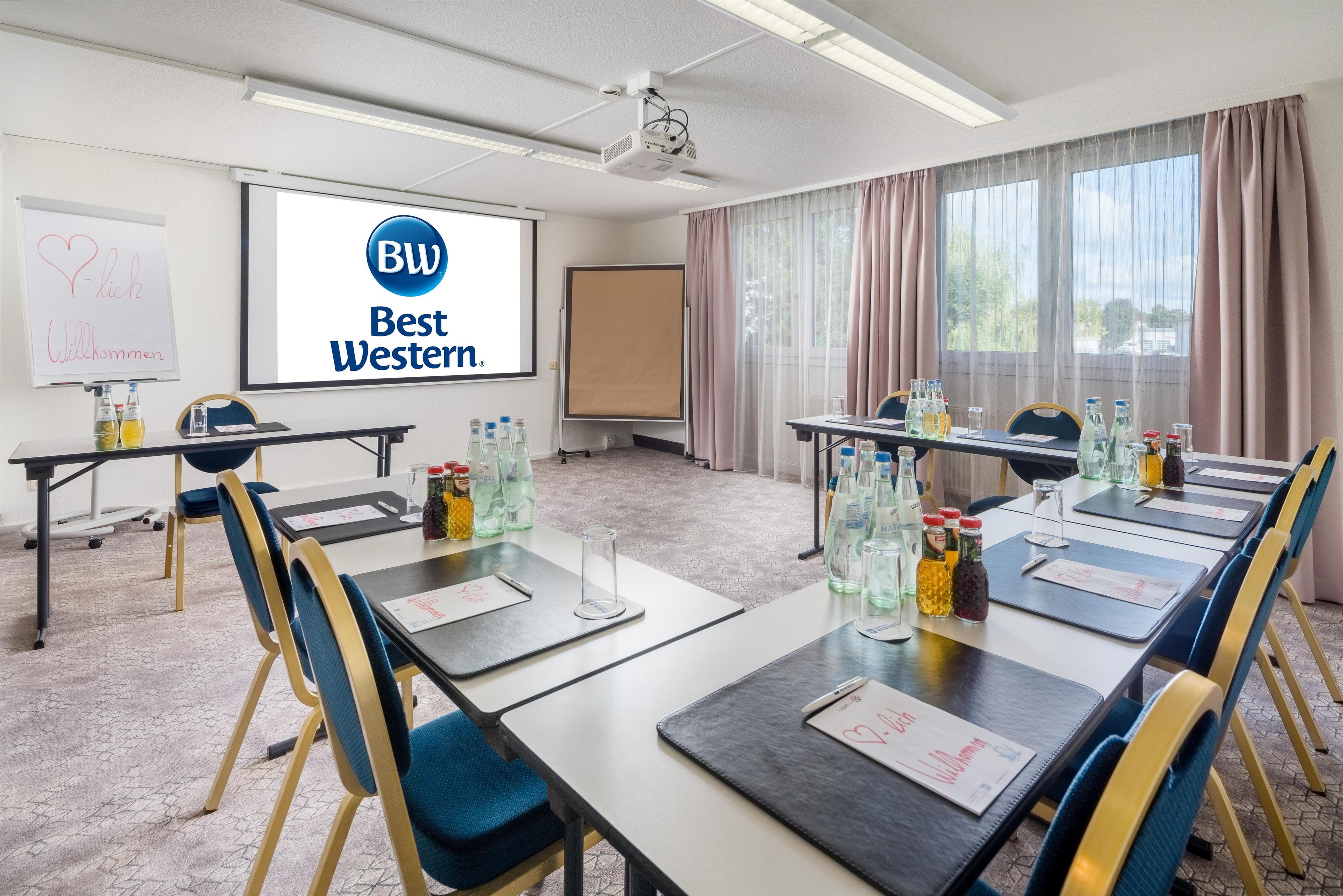 Ein Konferenzraum mit Projektionsschirm, der das Logo von 'BW Best Western' zeigt, Tische mit Wasserflaschen und einem Whiteboard mit Herzzeichnung und Text.