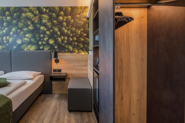 Un dormitorio compacto con cama, un banco, una mesita pequeña y un estante en la pared.