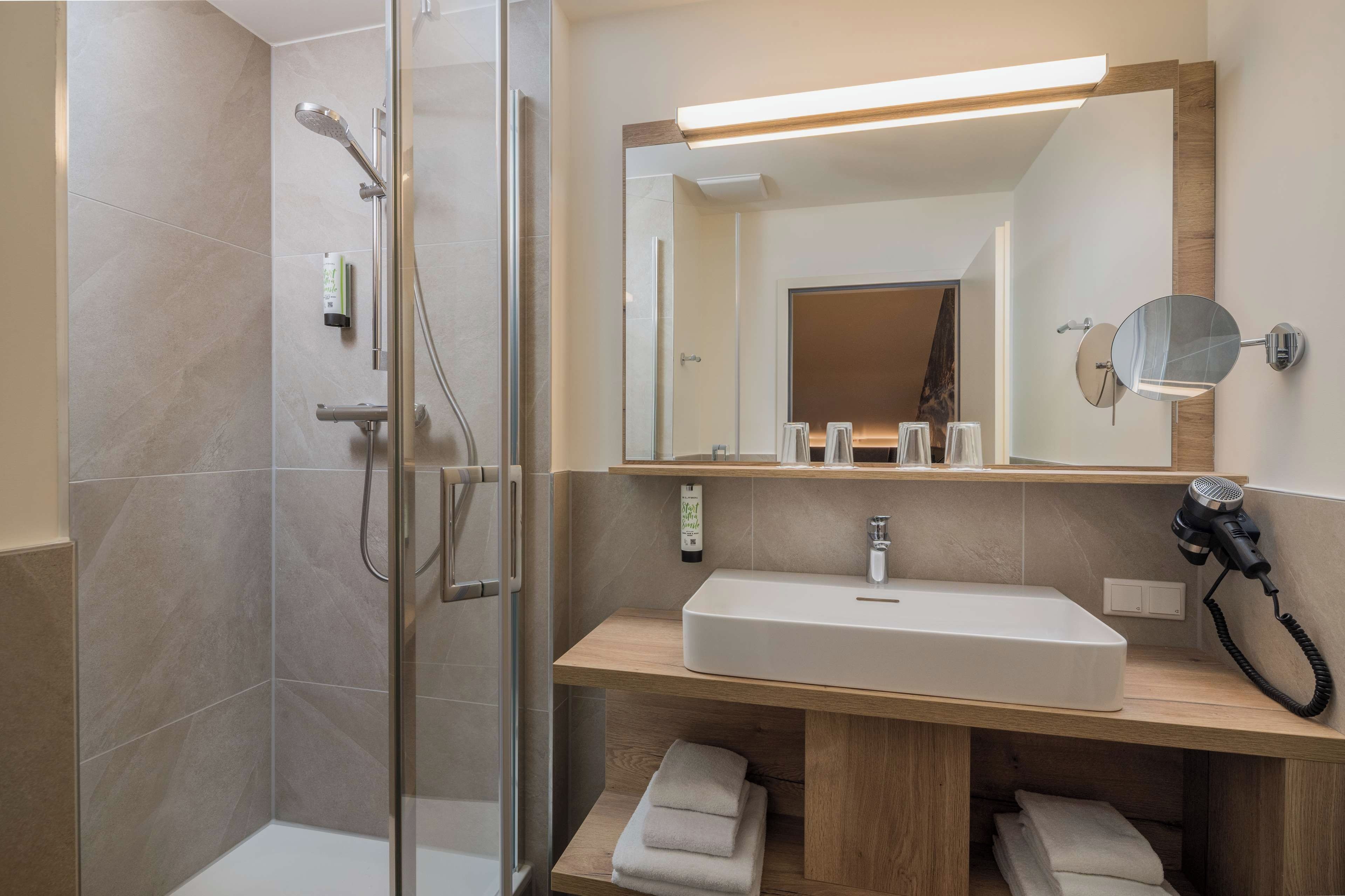 A modern bathroom with a glass-enclosed shower, a wooden vanity with a large mirror, and a hairdryer on the counter.