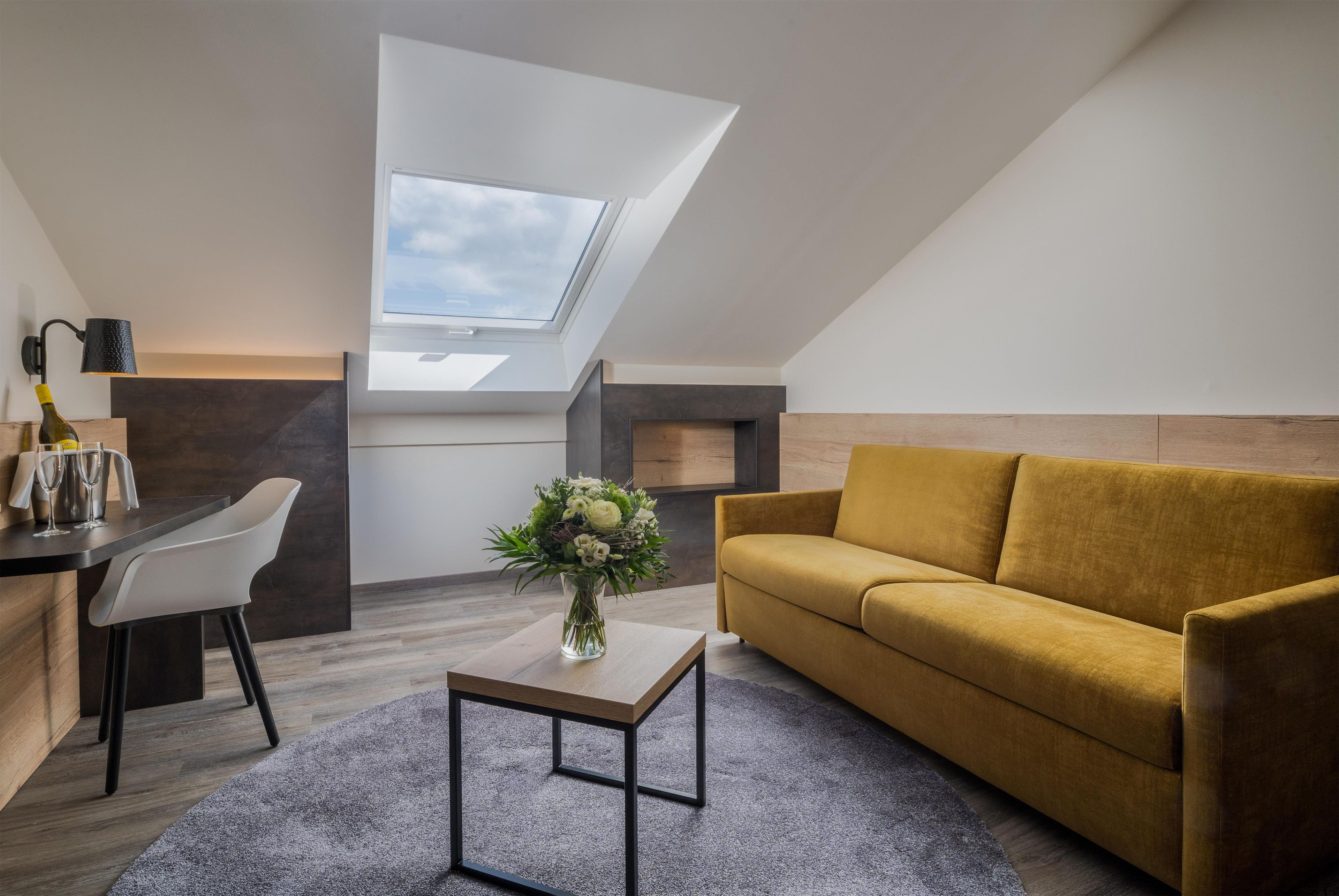 A modern loft apartment with a skylight, a sofa, a dining table with chairs, and a vase of flowers.