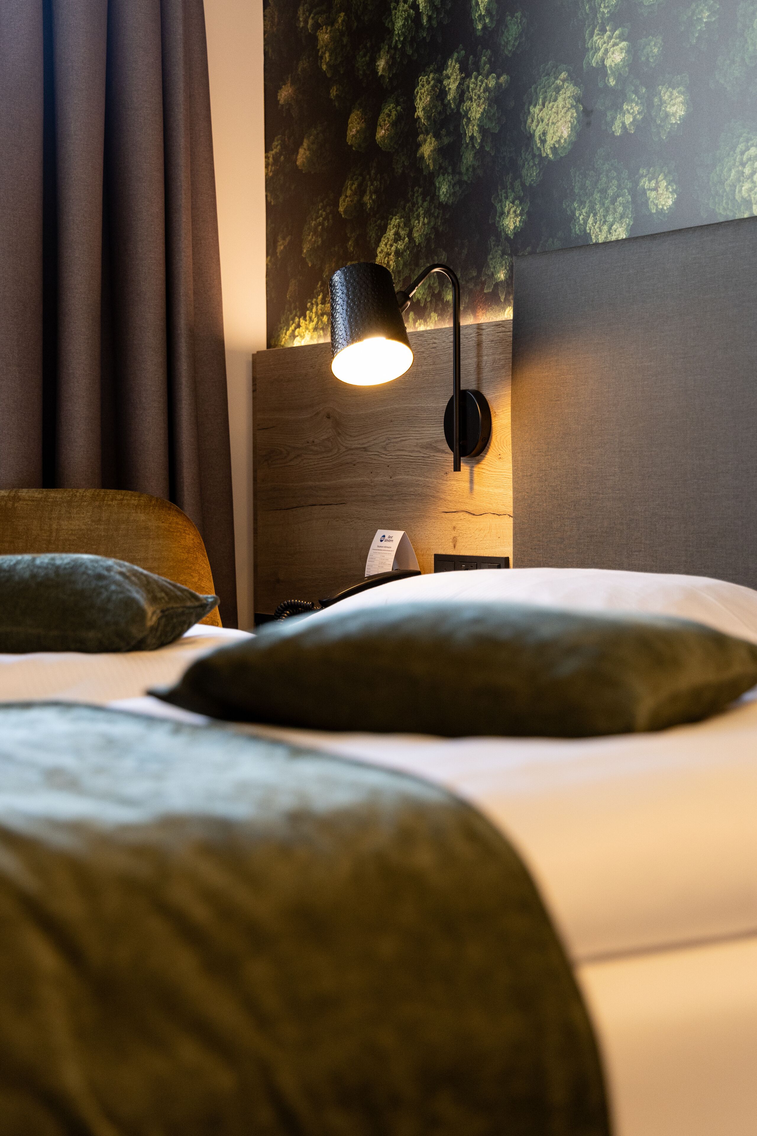 Ein ordentlich bezogenes Bett mit dunkelgrüner Bettwäsche, eine Wandlampe und eine strukturierte Wand dahinter.
