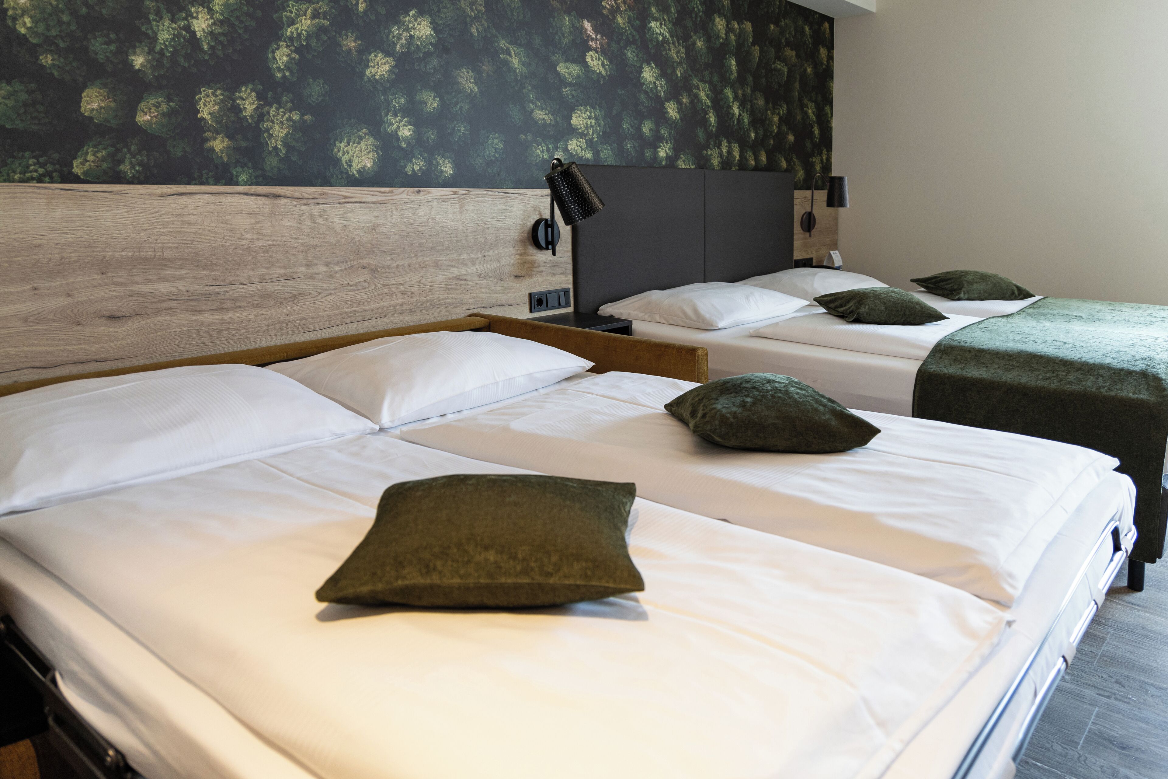 A hotel room with two beds, each with white linens and green pillows. There is a wooden headboard, a wall-mounted lamp, and a dark-colored wall with a leafy pattern.