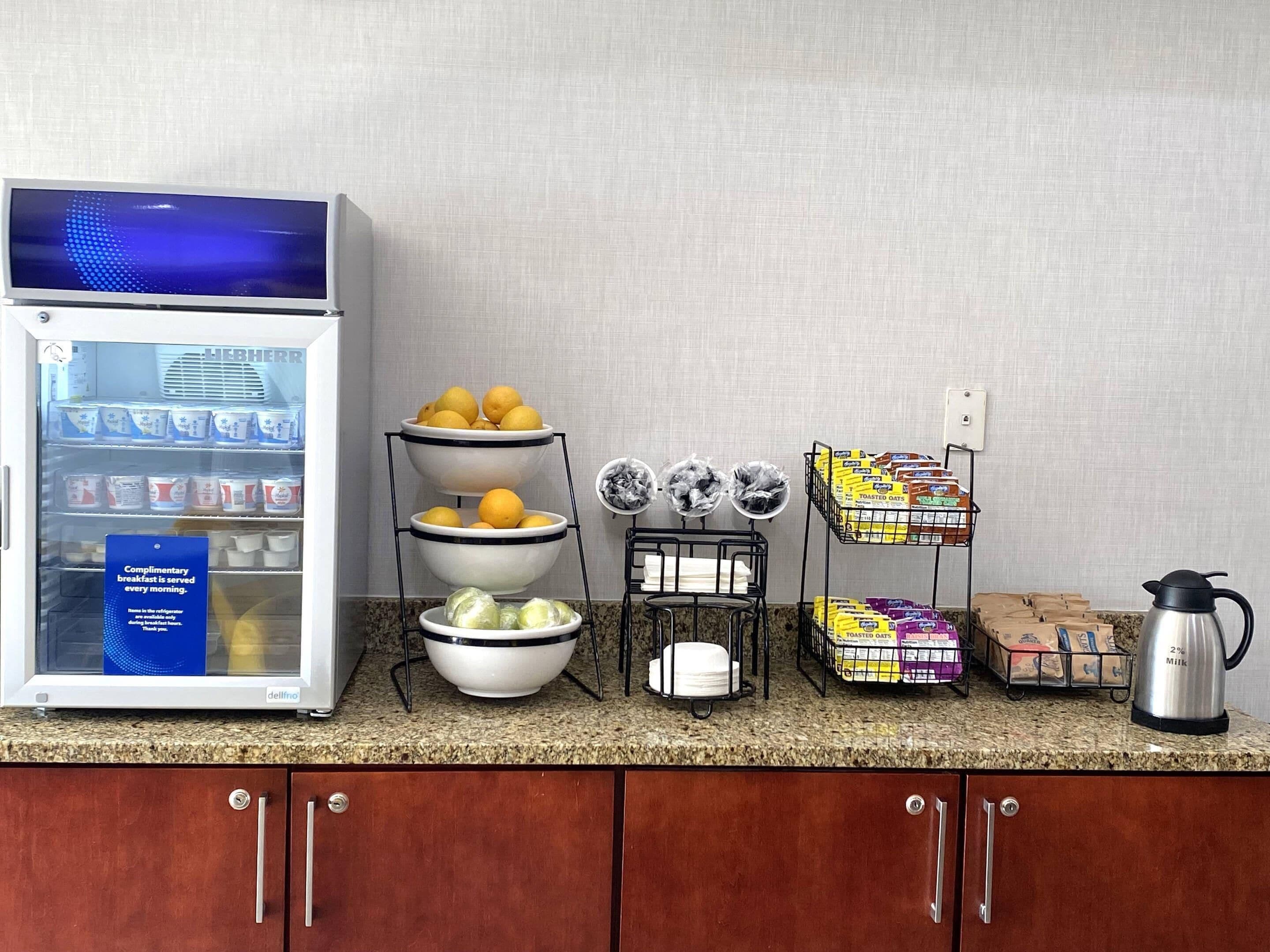 A breakfast bar with a refrigerator stocked with dairy products, a fruit bowl, a coffee maker, and a display of packaged goods.