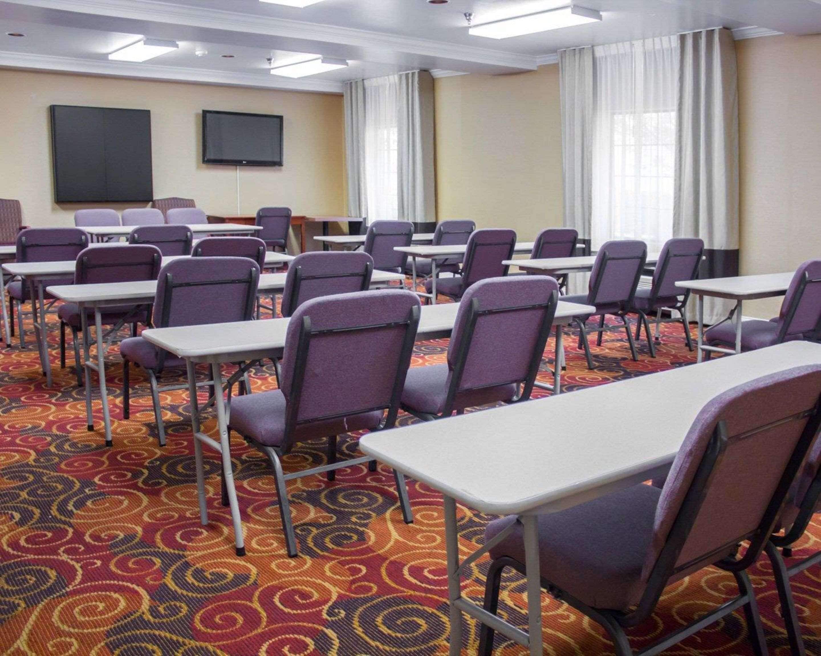 Un salón de conferencias con filas de sillas moradas y mesas blancas, una pantalla grande y alfombra estampada.