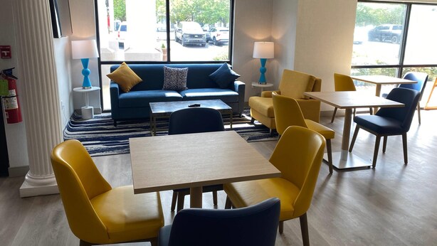 Een hotellobby met een blauwe bank, gele stoelen, een houten tafel en grote ramen met uitzicht op geparkeerde auto's.