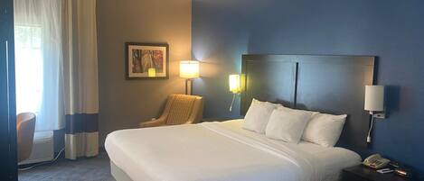 Una habitación de hotel con una cama grande, dos lámparas de noche, una silla, un cuadro en la pared y un teléfono en la mesita.