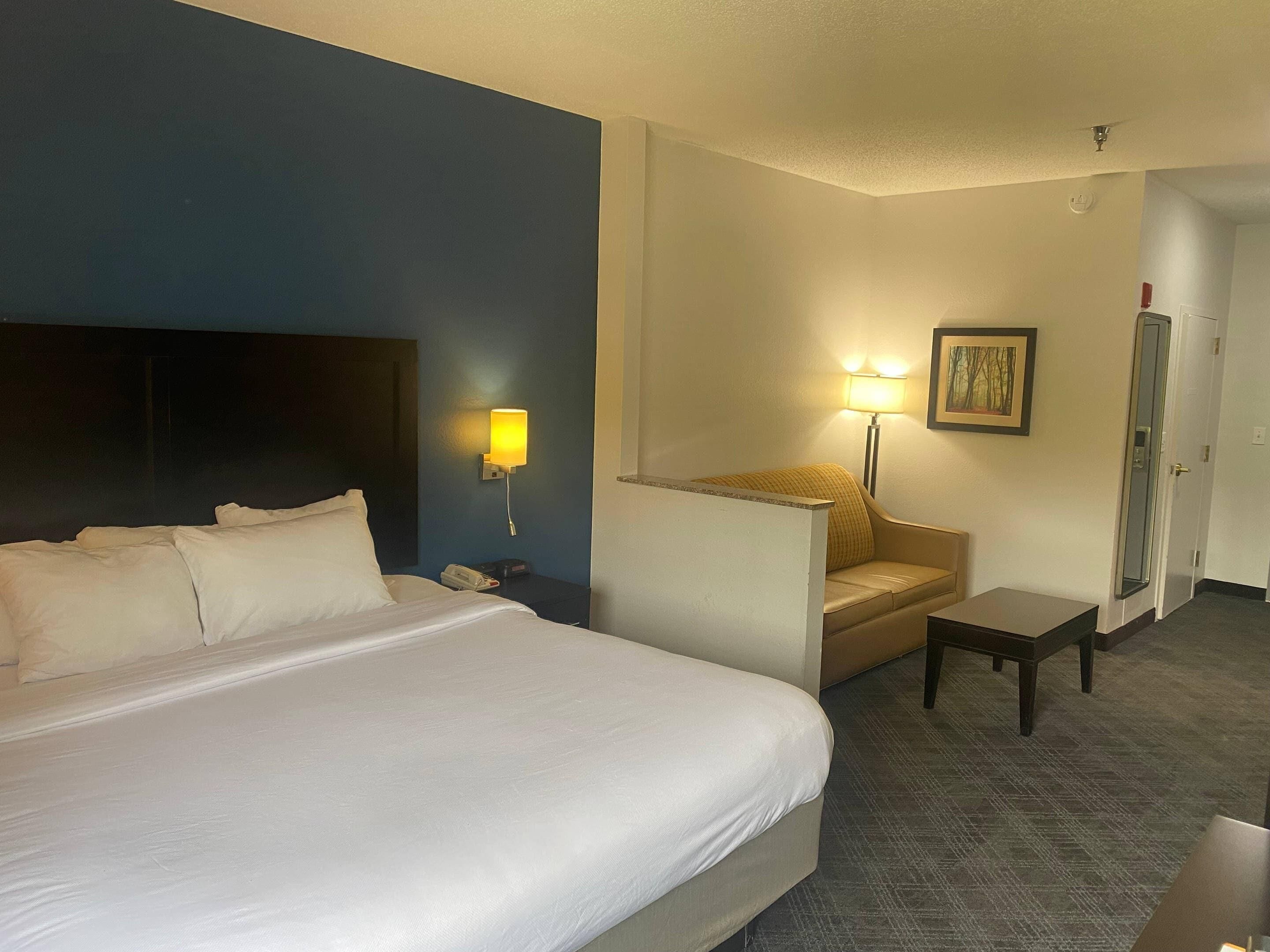 A hotel room with a large bed, a nightstand with a lamp, a small table, a sofa, and a painting on the wall.