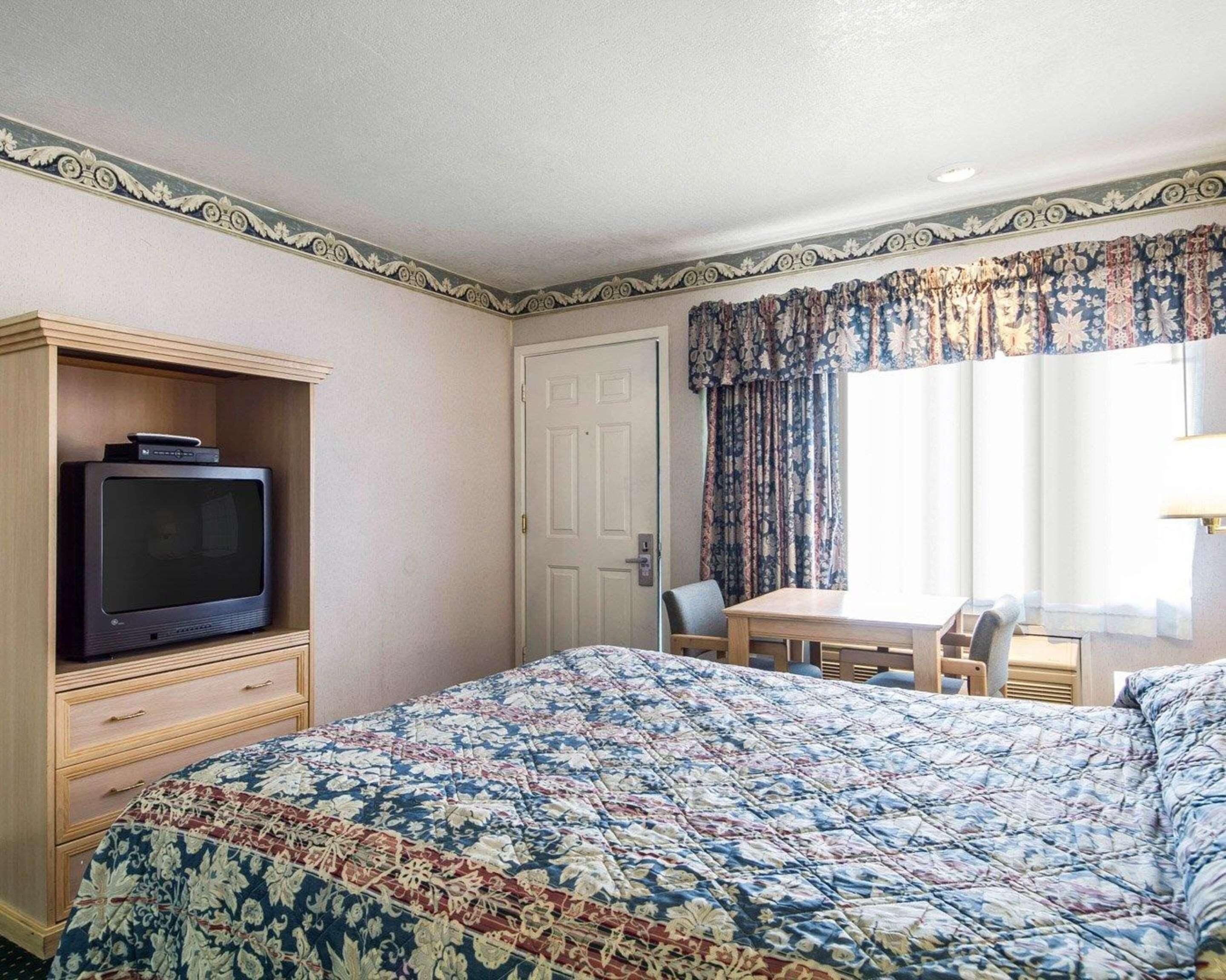 Une chambre d’hôtel avec un lit, un téléviseur posé sur un meuble en bois, une table à manger avec des chaises et une fenêtre avec des rideaux.