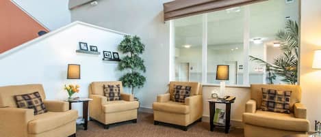 A modern living room with four beige armchairs, a coffee table, and a large window with a view of the outside. There are framed pictures on the wall and a potted plant.