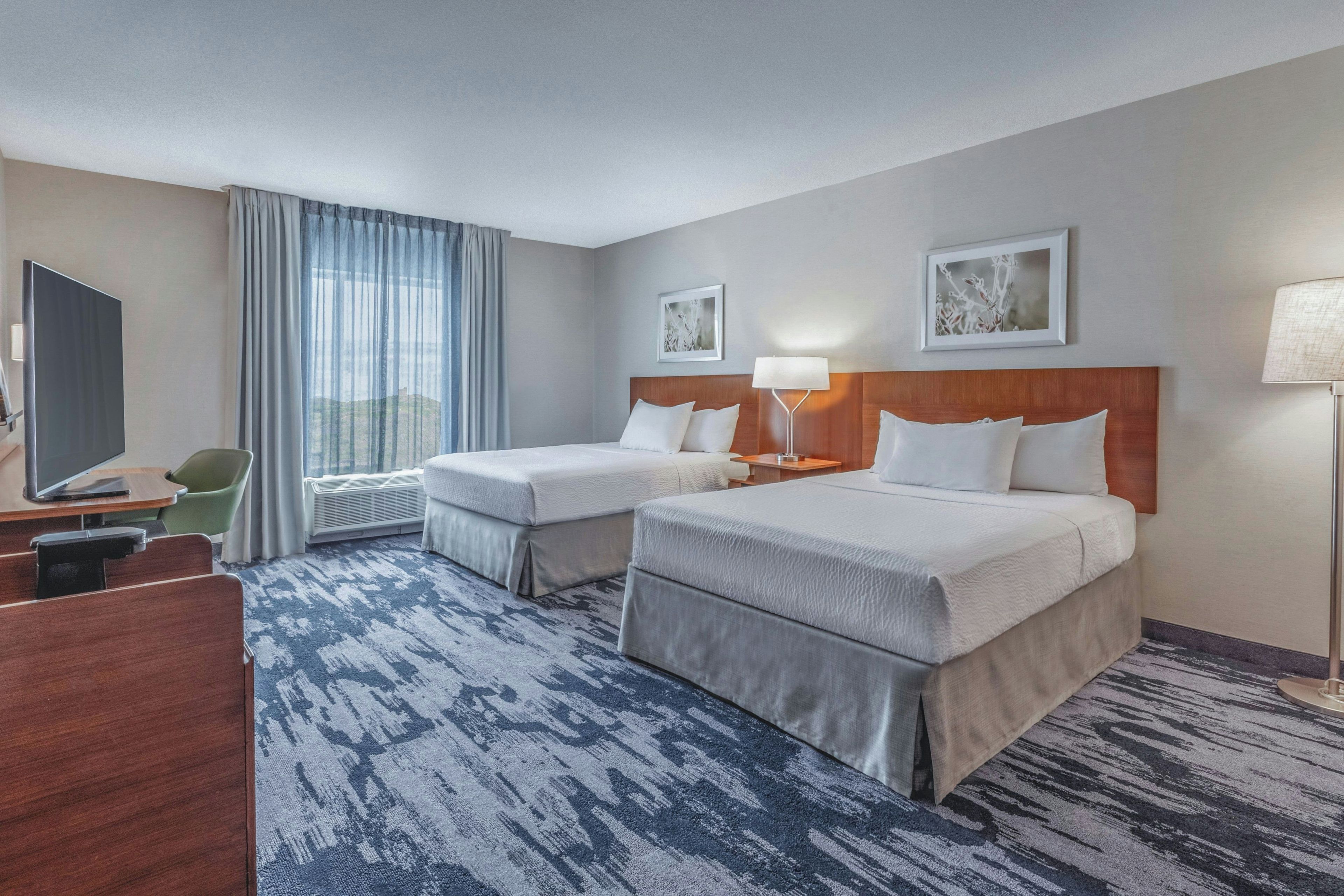 A hotel room with two beds, a desk, a chair, a TV, and a large window with curtains.
