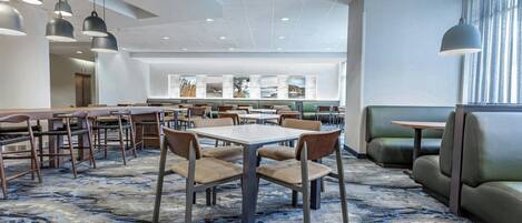 A modern hotel lobby with multiple seating areas, including tables and booths, and a patterned carpet.