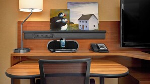 Habitación de hotel con un escritorio de madera, una lámpara, un cuadro con frailecillos y una casa, un televisor y un teléfono.