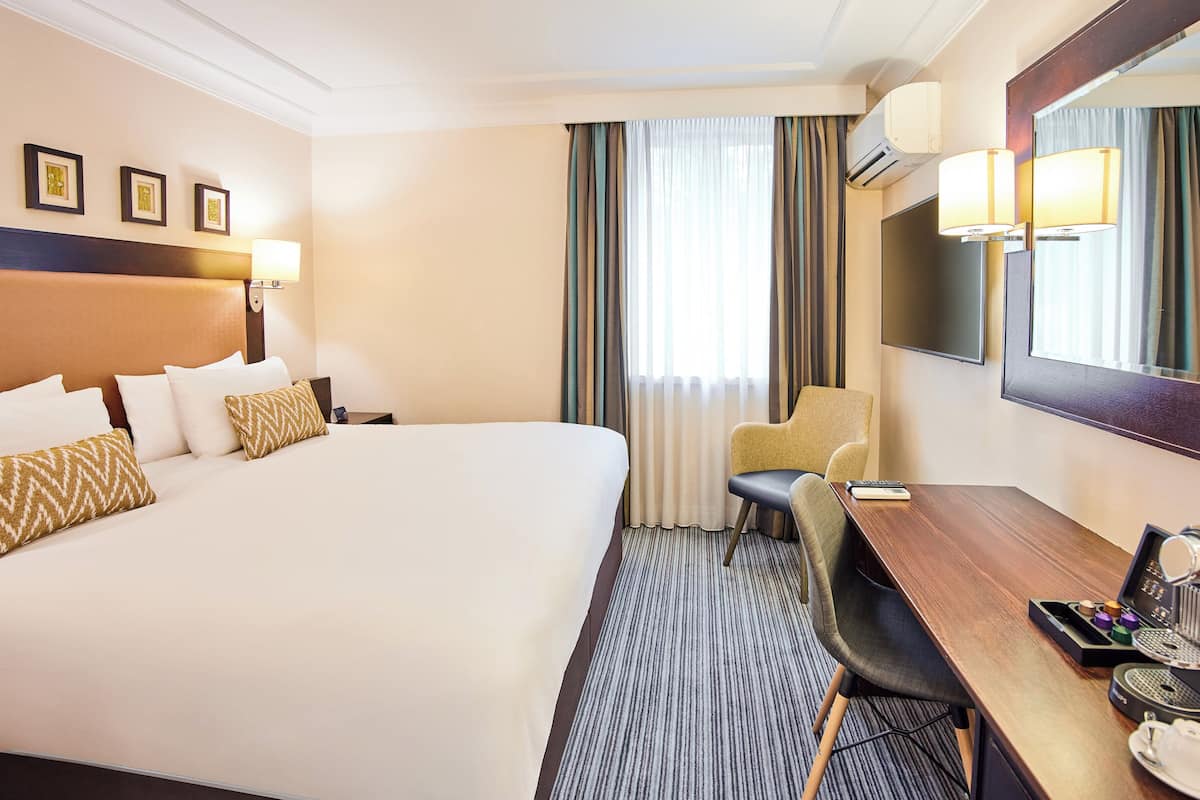 A hotel room with a large bed, a desk with a chair, a coffee machine, and a TV.