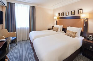 A hotel room with two beds, a desk, a chair, a TV, and a window with curtains. - Thistle London Heathrow Terminal 5 (West Drayton)