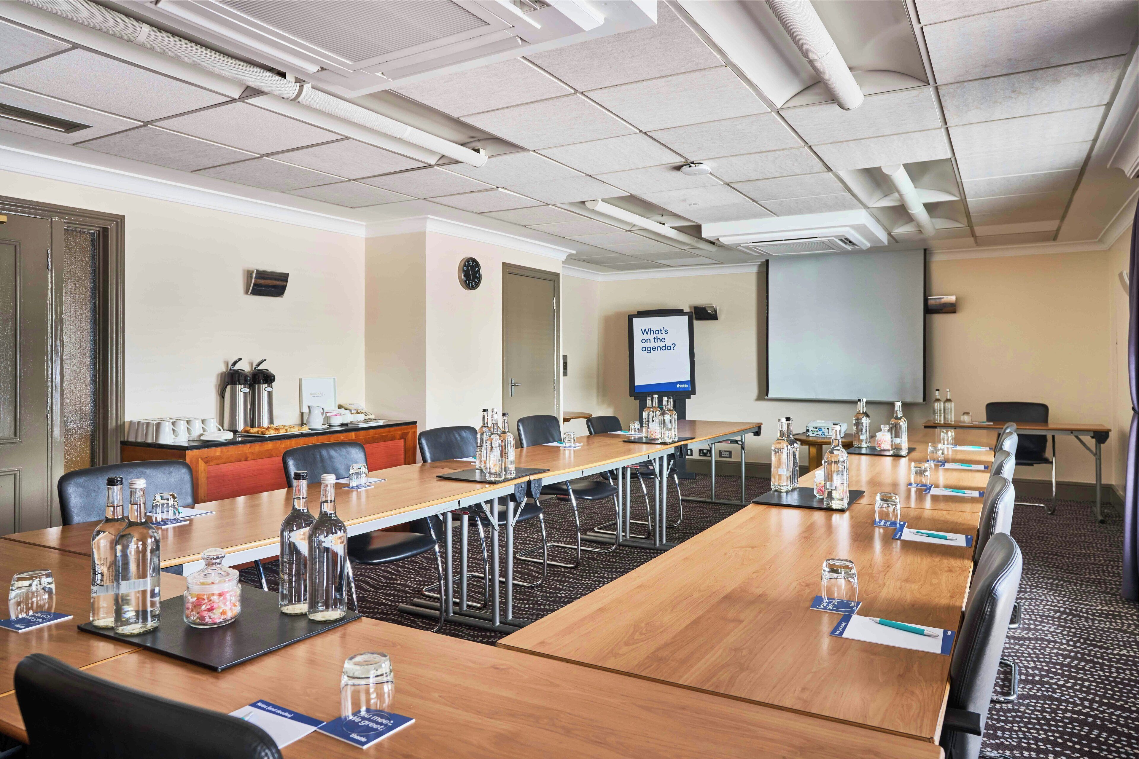 A conference room with a long table, chairs, a projector screen, and a coffee station.