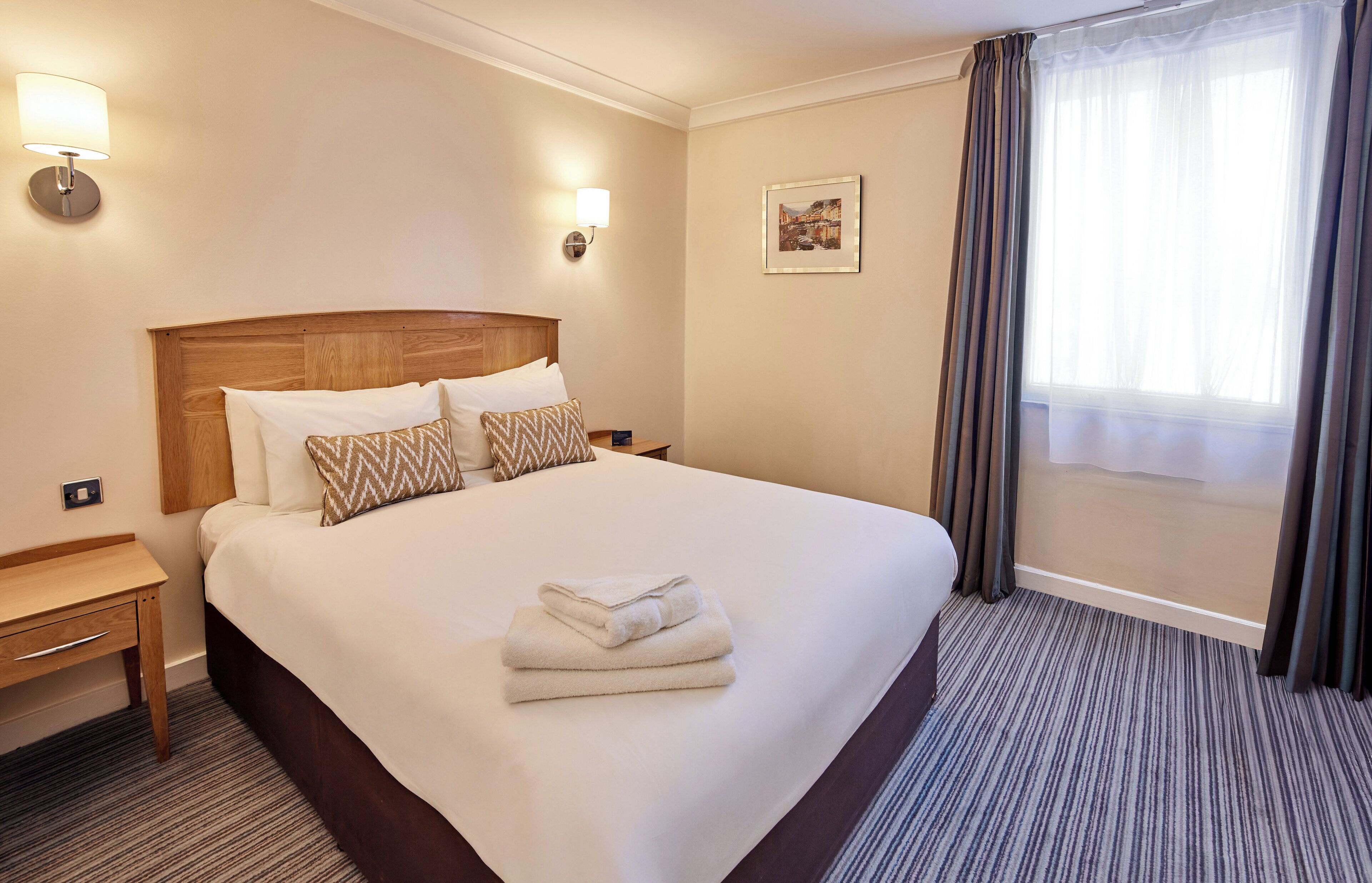 A hotel room with a large bed, two bedside tables, a nightstand, a lamp, a framed picture on the wall, and a window with curtains.