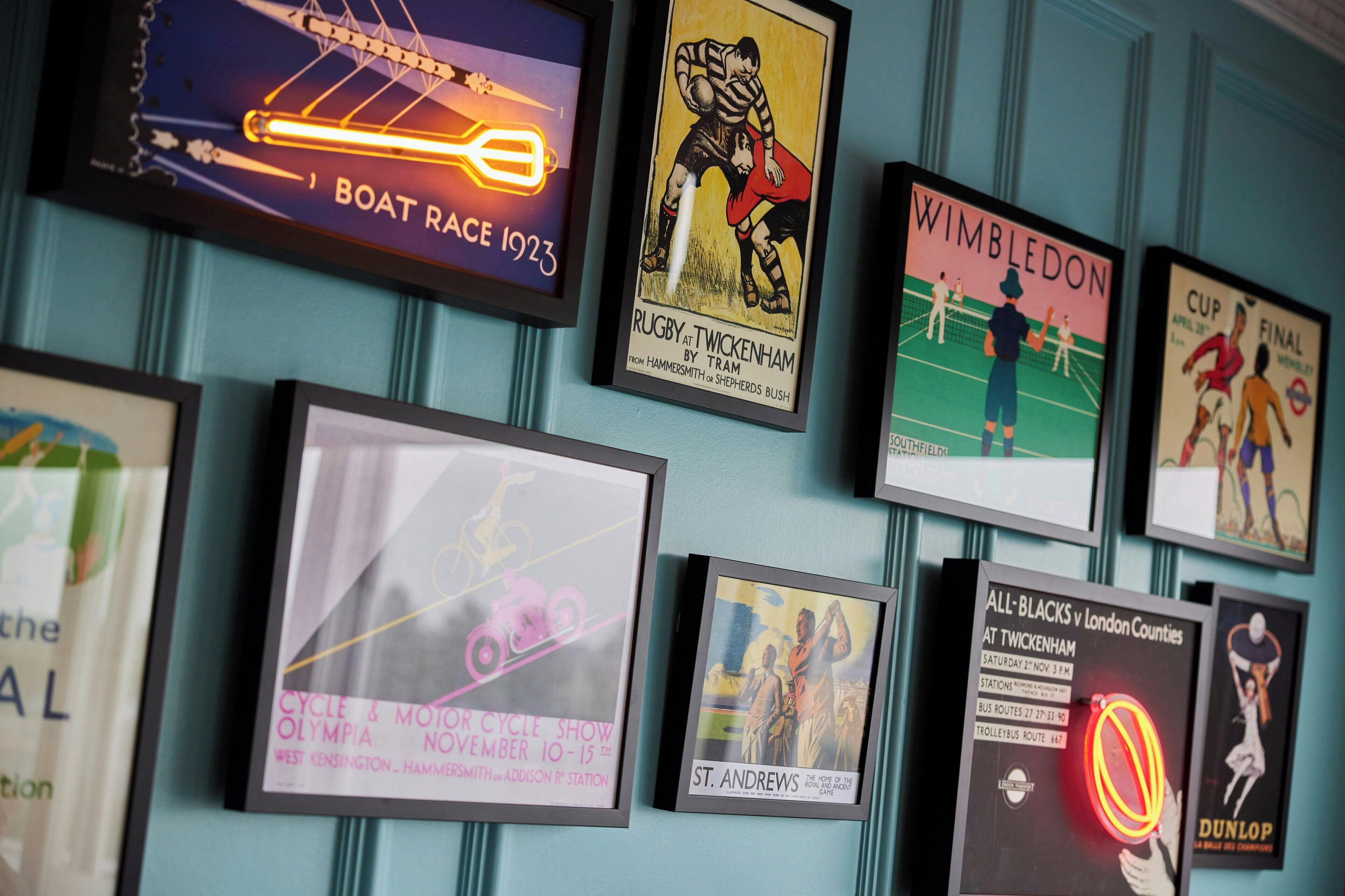 A wall with various framed posters, including a boat race, rugby, Wimbledon, and a cycle show.