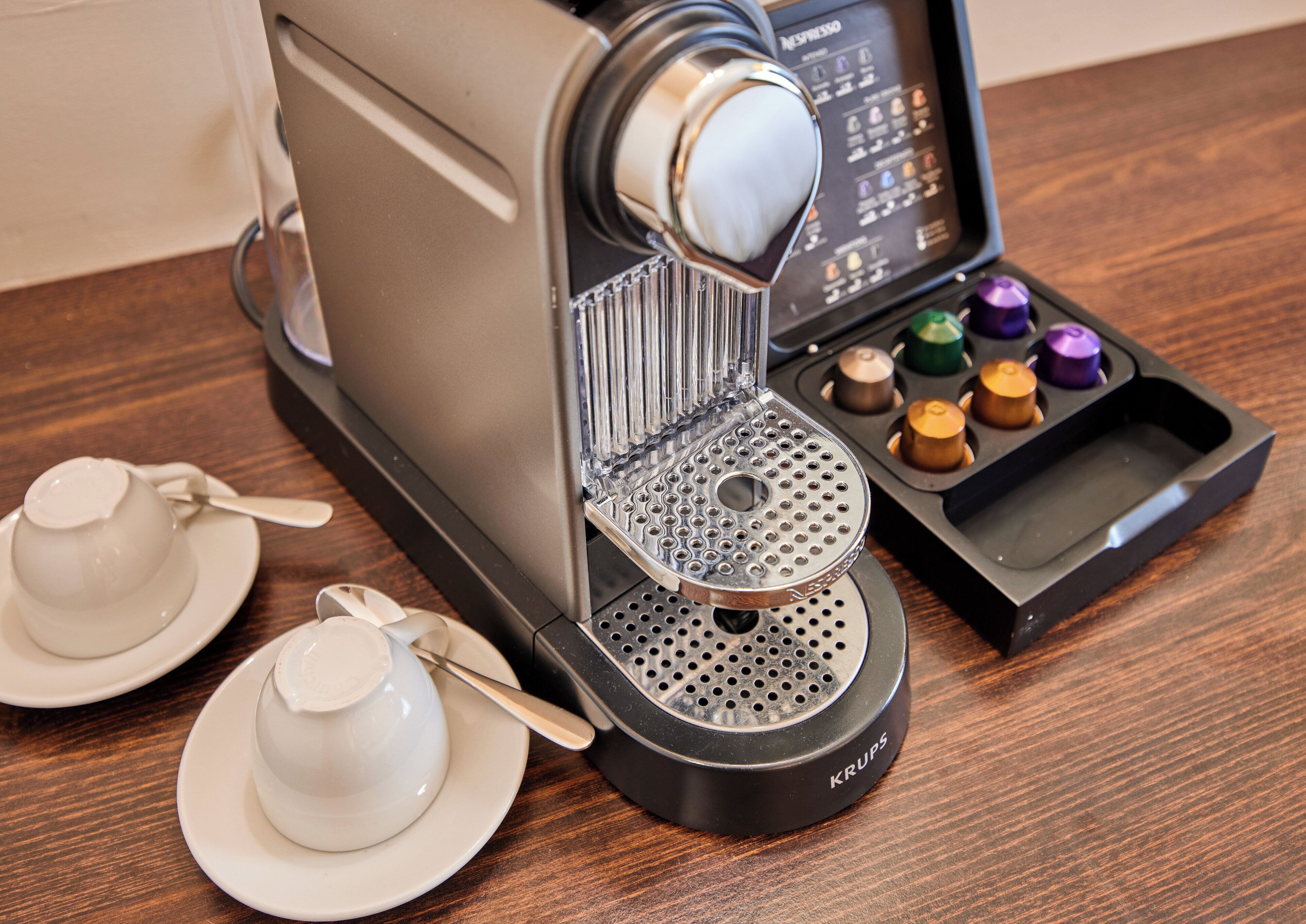 A Krups coffee machine with a drip tray, two white cups, and a spoon on a wooden table.