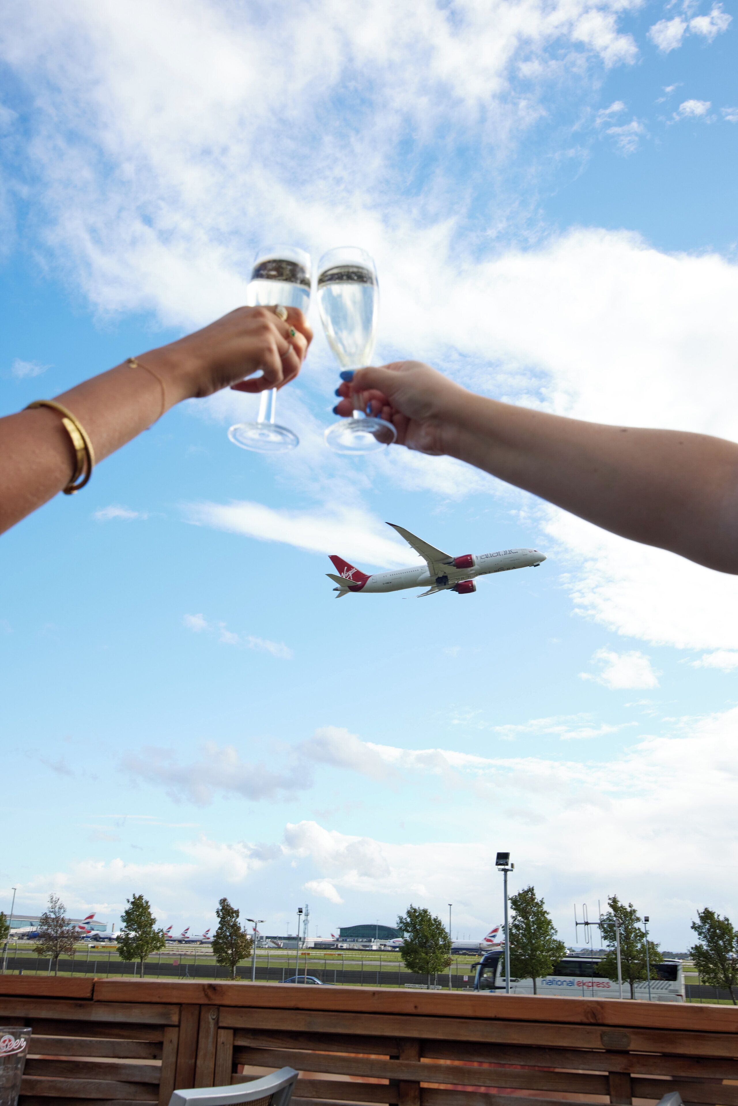 Two hands holding champagne glasses, an airplane in the background, and a view of a runway with airport buildings.