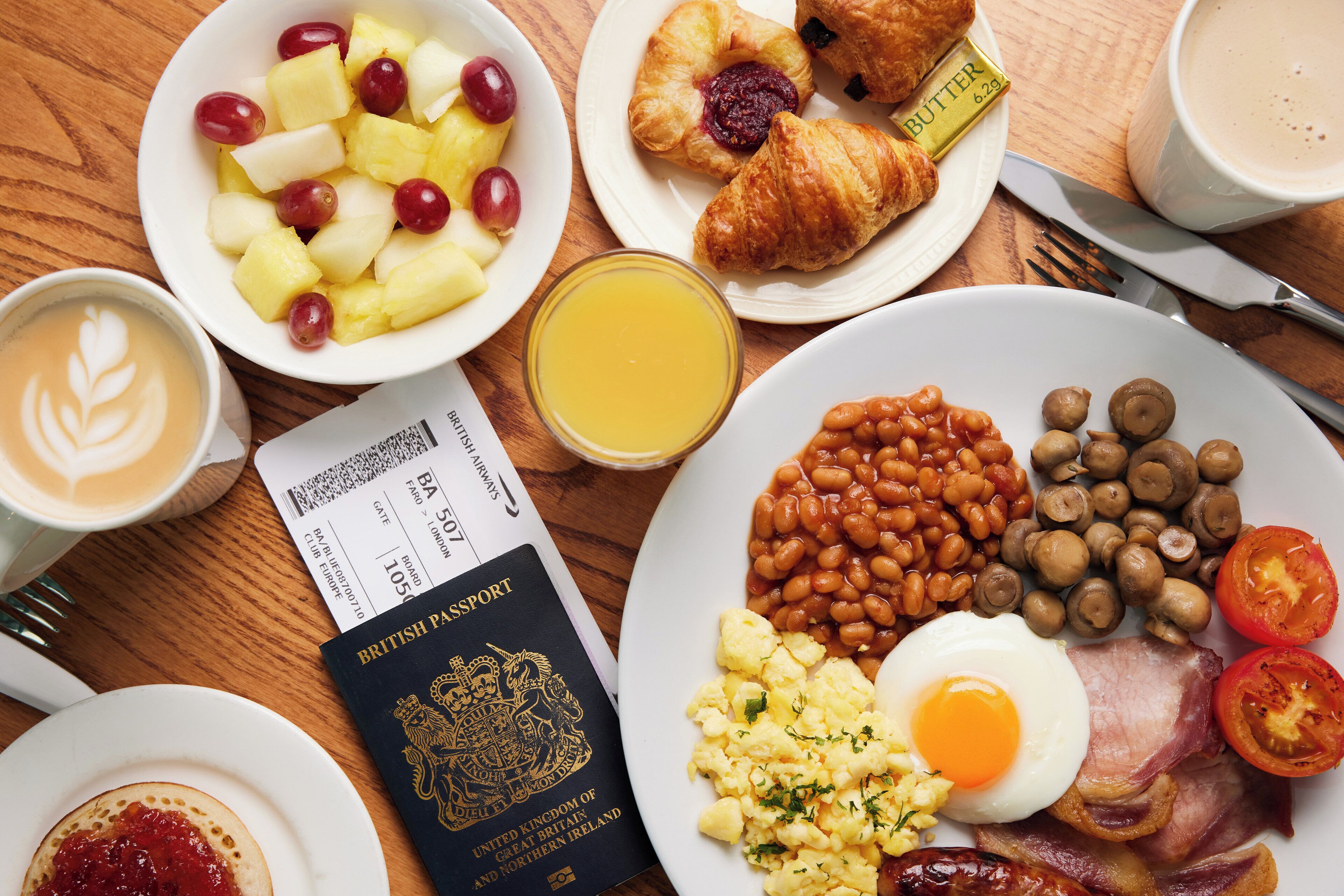 A breakfast table with a passport, a croissant, a plate of beans, mushrooms, tomatoes, and a plate of scrambled eggs and bacon.