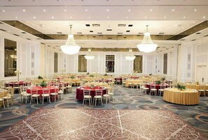 Um amplo salão de banquetes com mesas redondas, lustres e um tapete estampado.