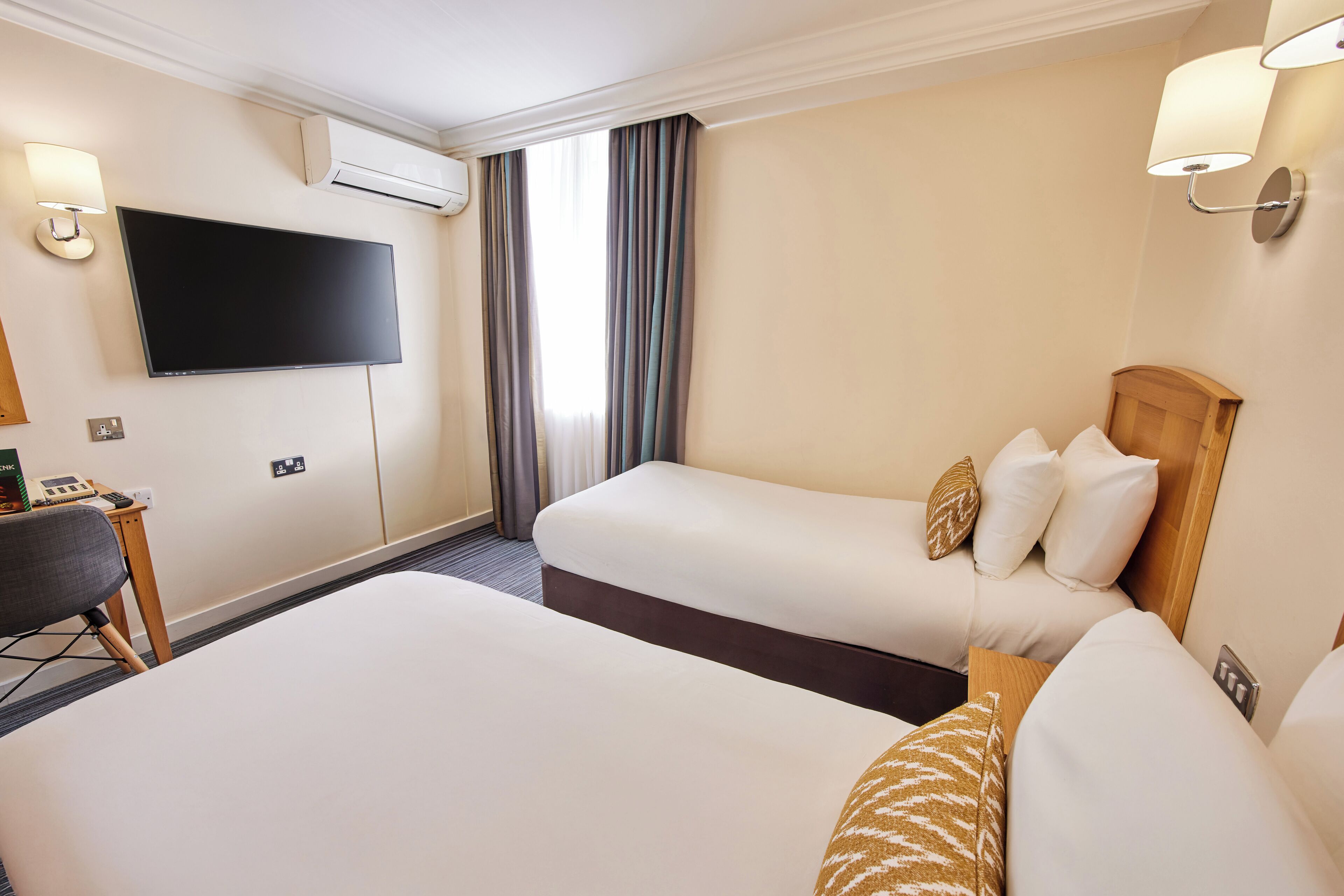 A hotel room with two beds, a flat-screen TV mounted on the wall, a desk with a chair, and a window with curtains.