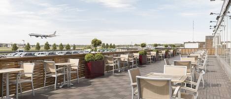 A rooftop terrace with outdoor seating, a view of an airport, and parked cars in the background.