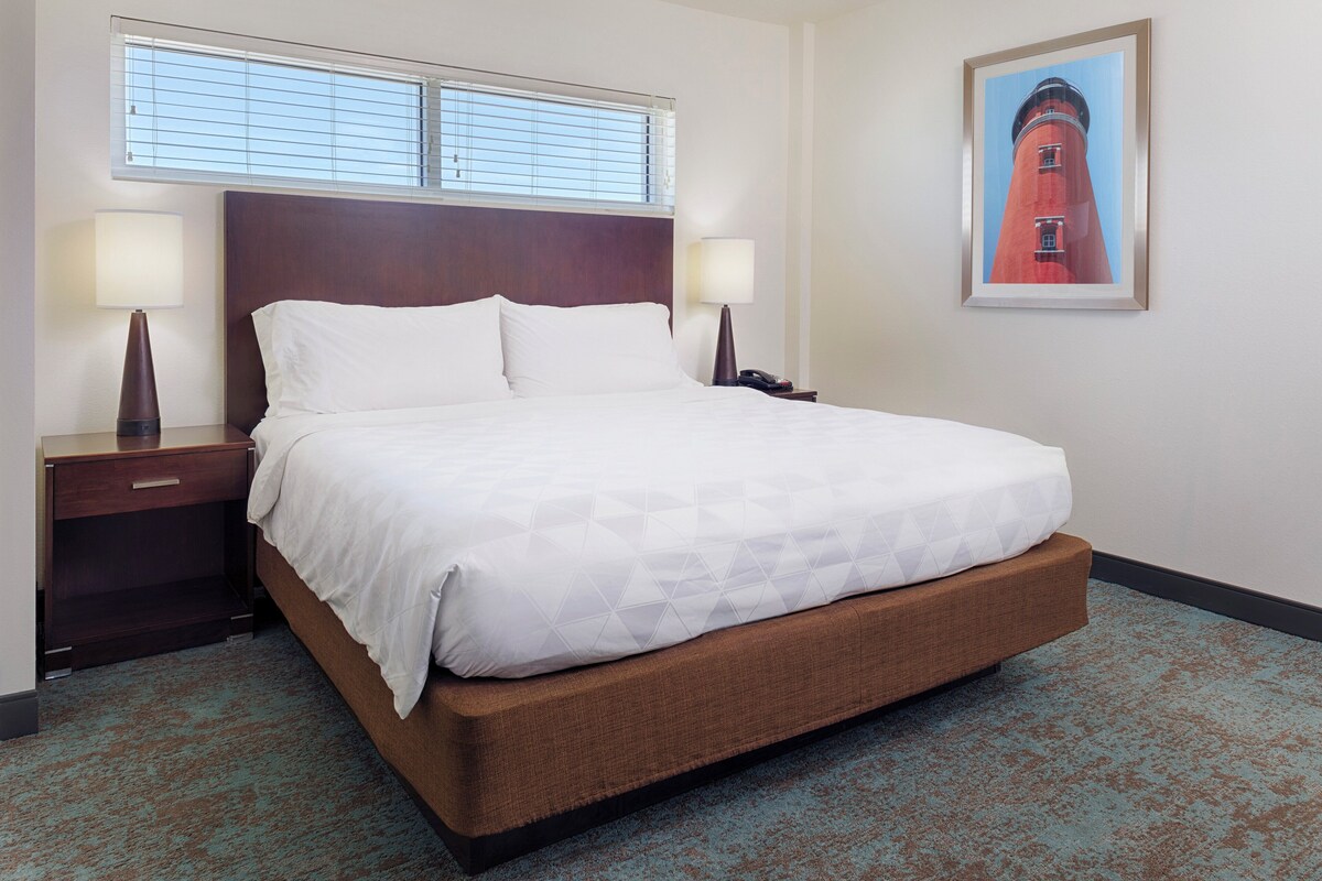 A hotel room with a large bed, two bedside tables with lamps, a window with blinds, and a framed picture on the wall.