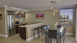 A modern kitchen with a built-in refrigerator, microwave, and oven. There is a dining area with a glass table and chairs, and a window with blinds.
