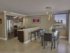 A modern kitchen with a built-in refrigerator, microwave, and oven. There is a dining area with a glass table and chairs, and a window with blinds. - Holiday Inn Resort Daytona Beach Oceanfront by IHG (Daytona Beach)