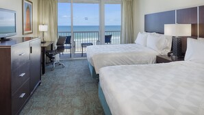 A hotel room with two beds, a desk with a chair, a television, and a view of the ocean through large windows.