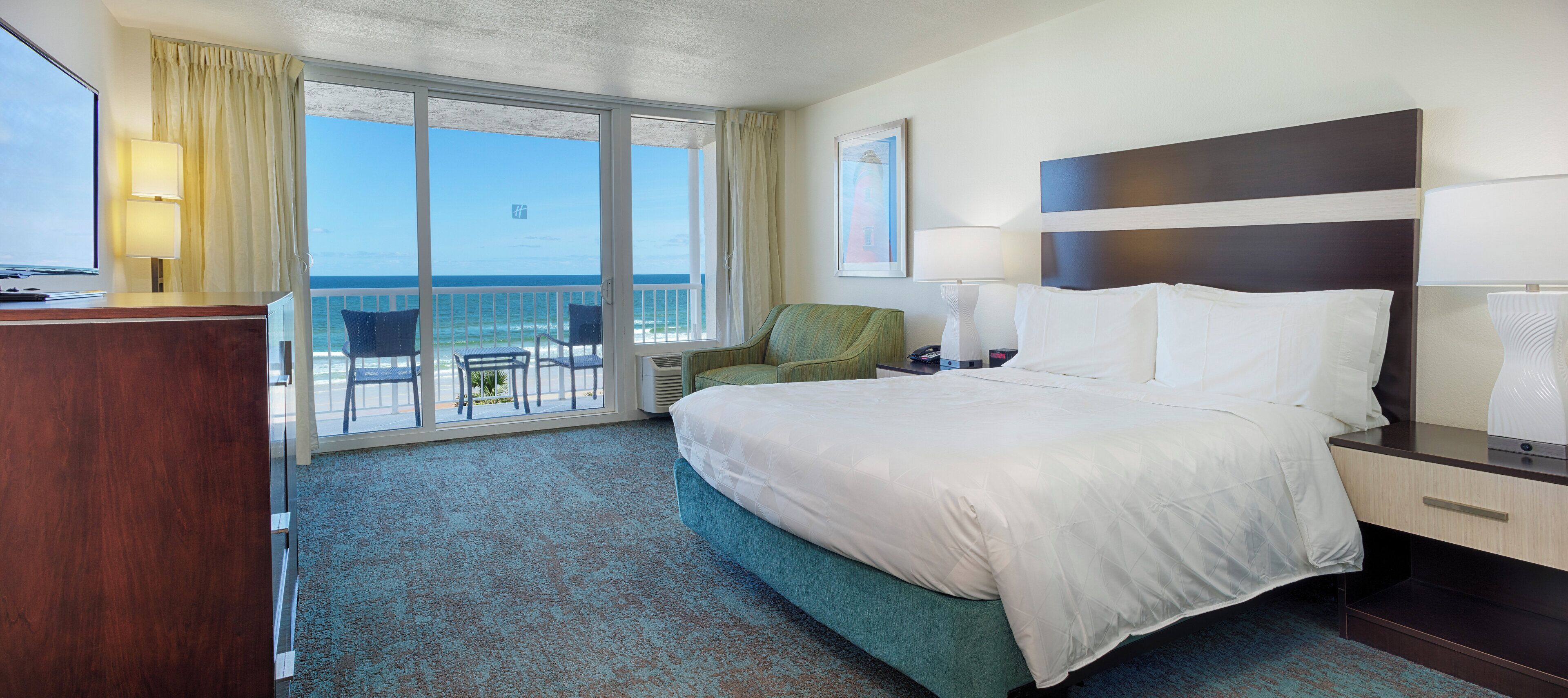A hotel room with a bed, a desk, a chair, a lamp, and a view of the ocean.