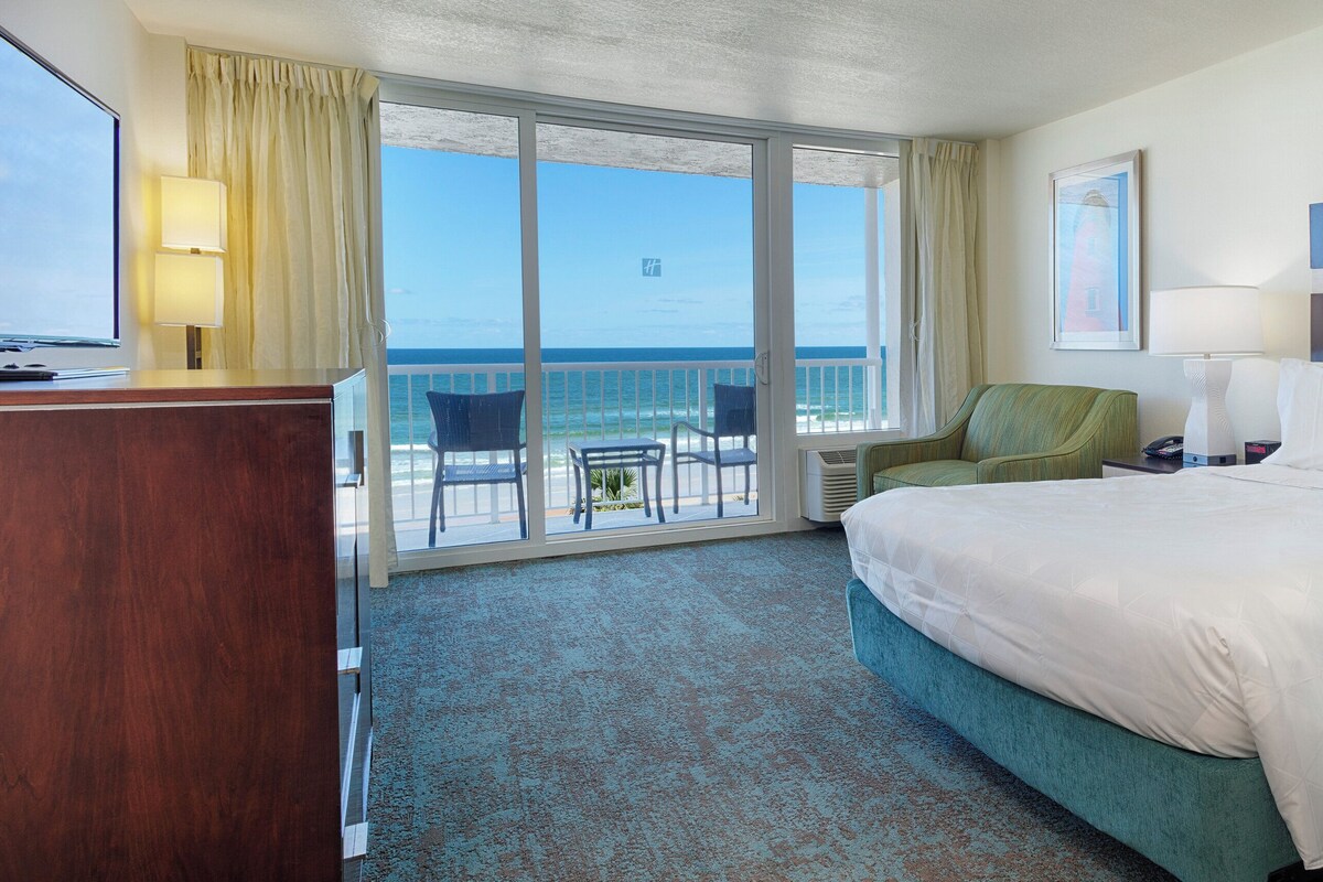 A hotel room with a bed, a desk, a chair, a lamp, and a view of the ocean.