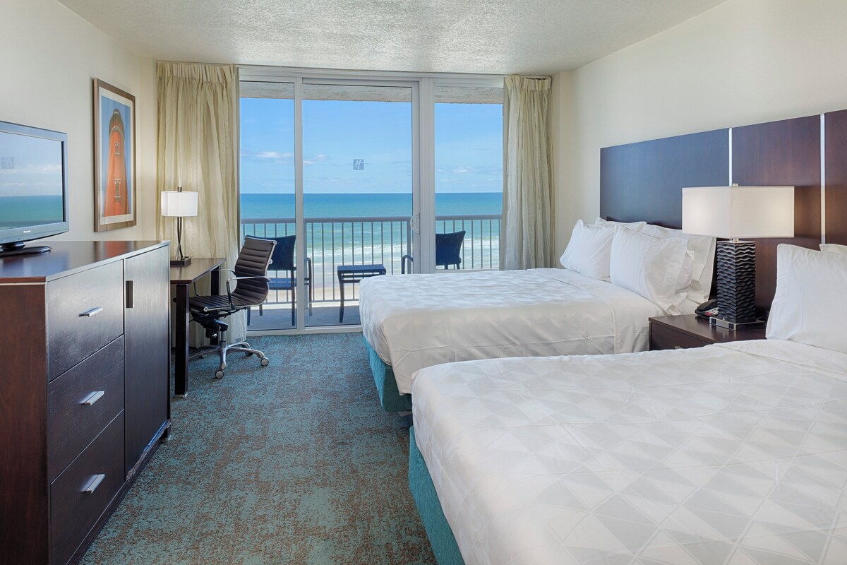 A hotel room with two beds, a desk with a chair, a TV, and a view of the ocean through large windows.