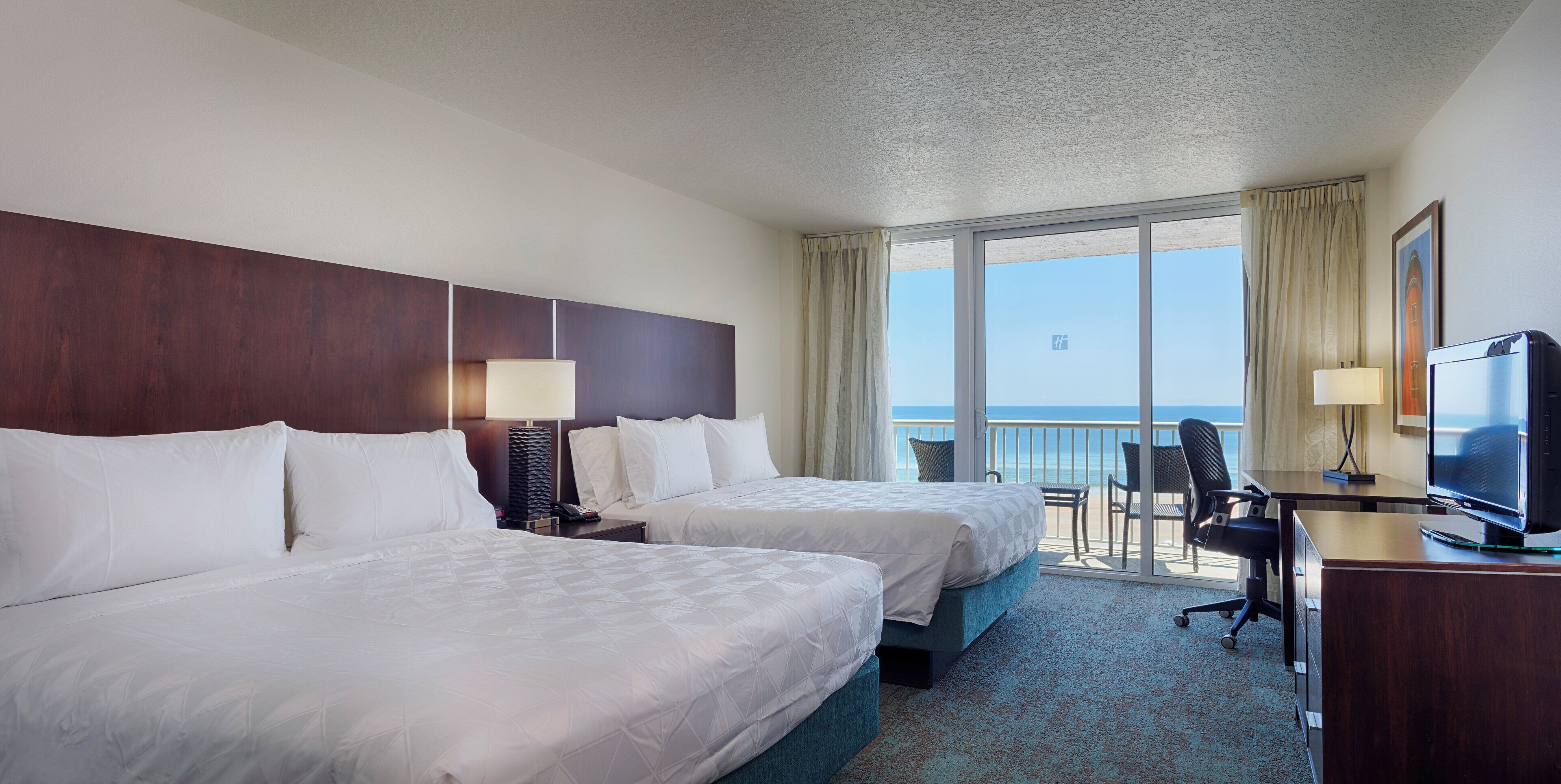 A hotel room with two beds, a desk with a television, and a balcony with a view of the ocean.