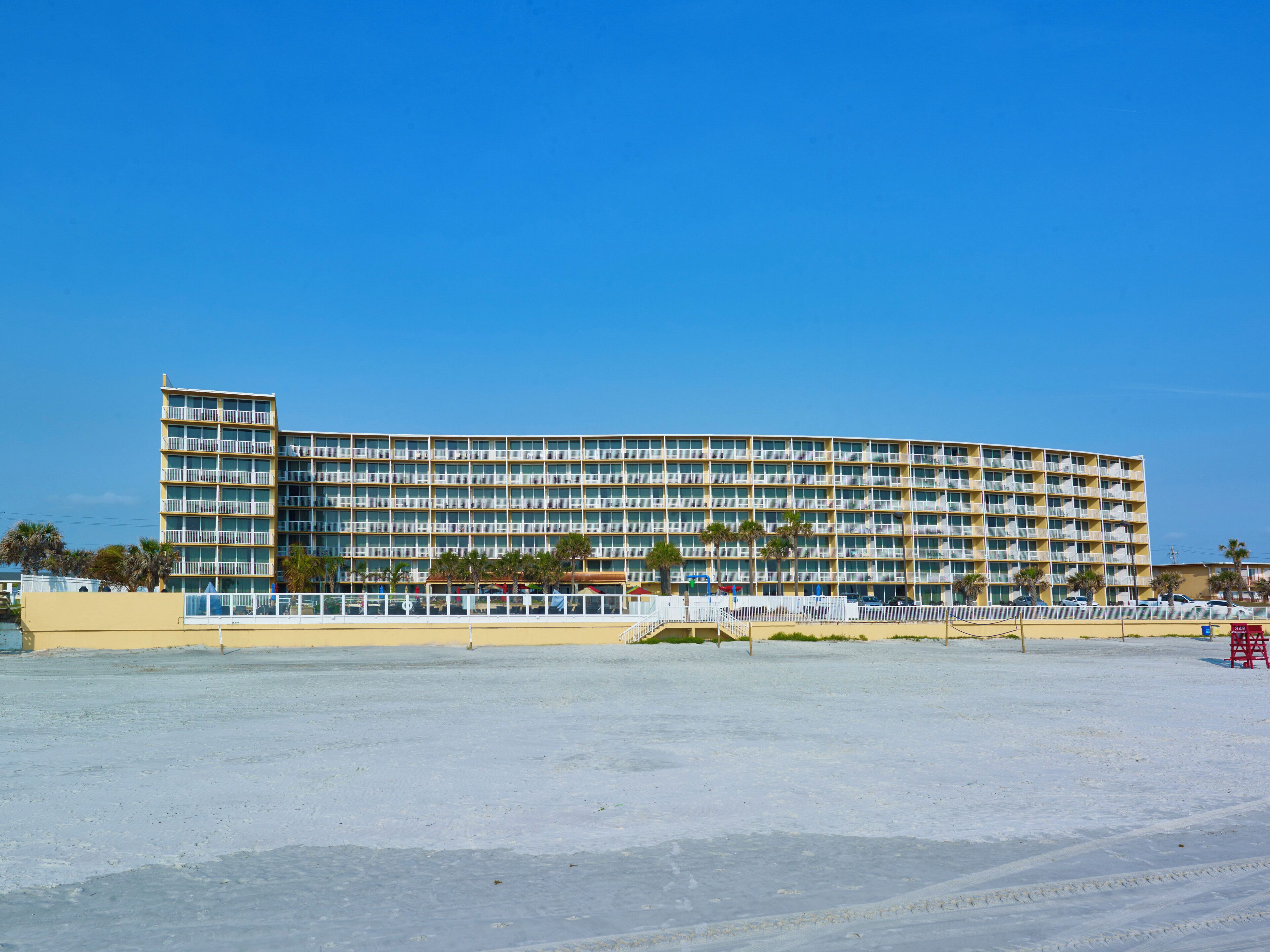 Un hotel de varios pisos con fachada plana, balcones y una playa de arena.