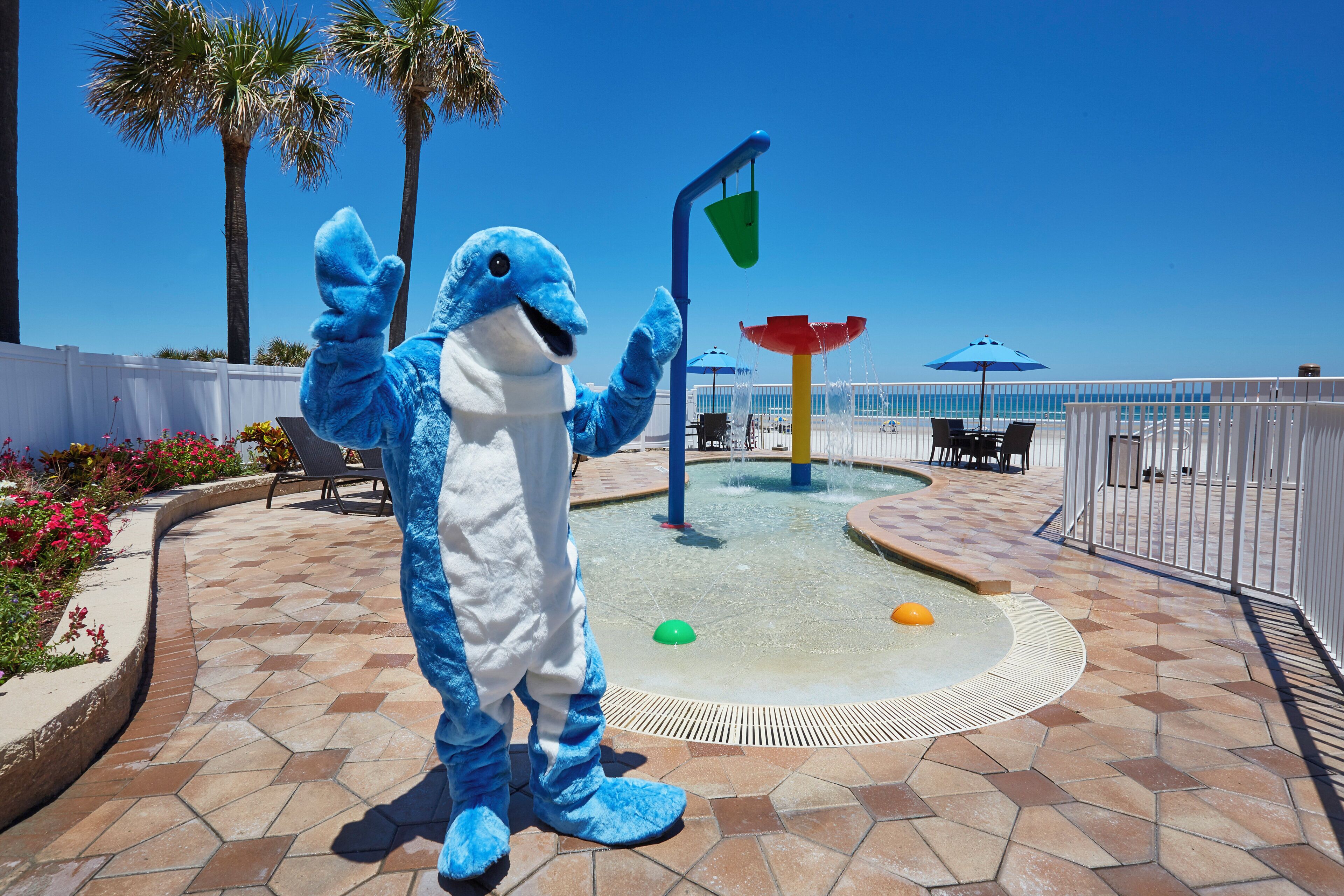 Um mascote com um fato azul em um pátio ladrilhado, com piscina, palmeiras e guarda-sóis de praia.