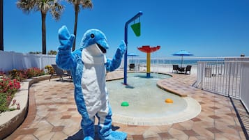 Um mascote com um fato azul em um pátio ladrilhado, com piscina, palmeiras e guarda-sóis de praia.