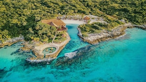 An aerial view of a resort with a central circular pool, surrounded by palm trees and a beach area.