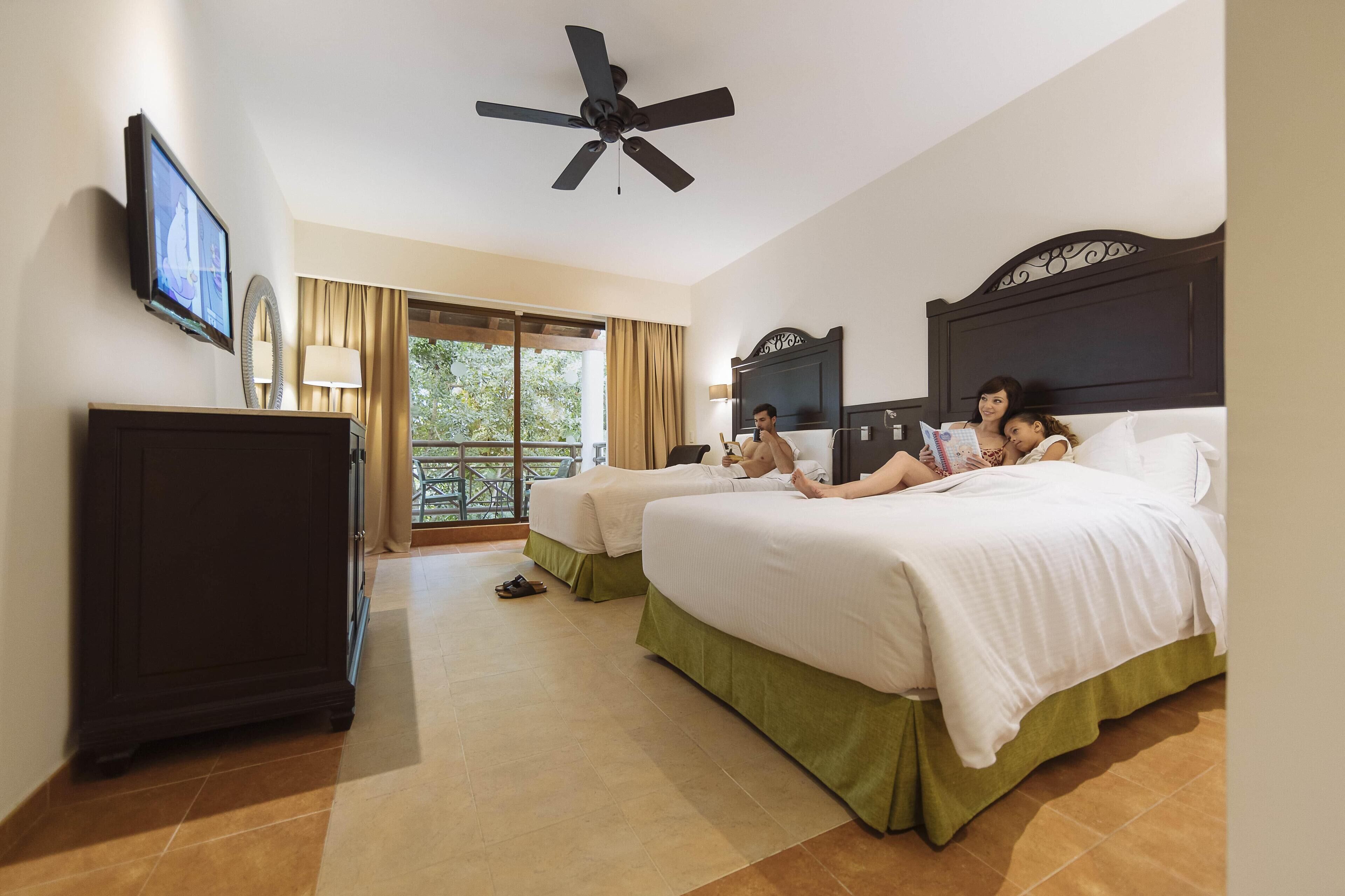 A hotel room with two beds, a ceiling fan, a TV, and a view of the outdoors.