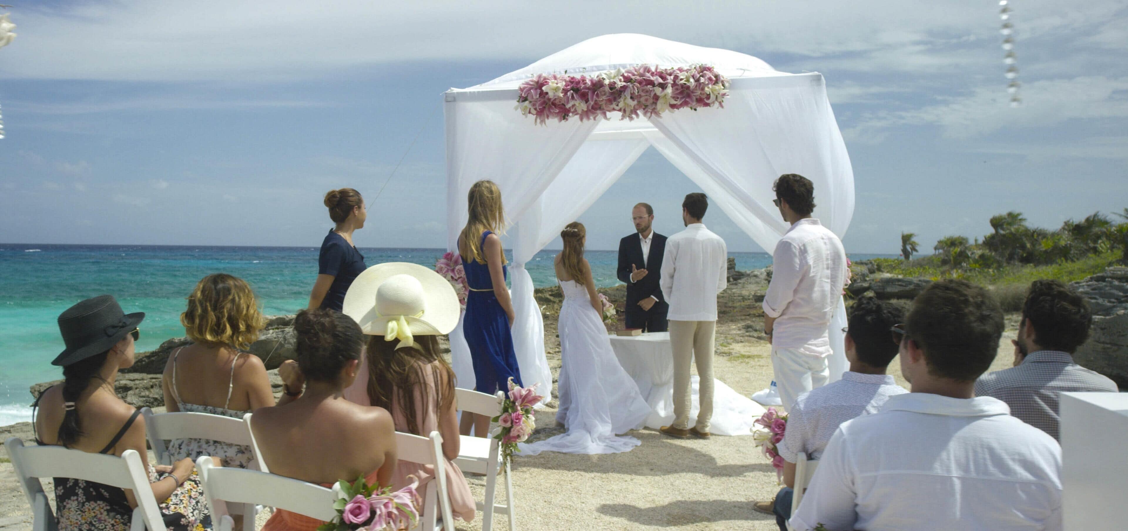 Een huwelijksceremonie op het strand, met een witte luifel en bloemversiering.
