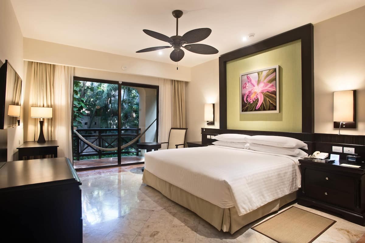 Ein Hotelzimmer mit einem großen Bett, einem Deckenventilator, einem Schreibtisch, einem Sessel, einem Nachttisch, einer Lampe, einem Bild und Blick ins Freie.