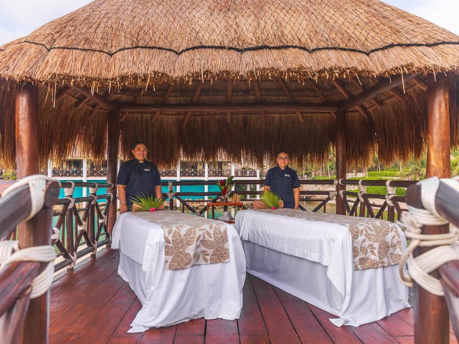 A thatched-roof gazebo with two beds, wooden railings, and a view of a pool and greenery.